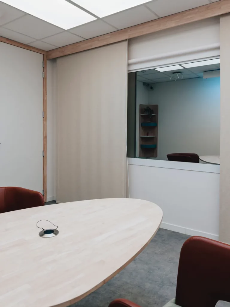 Videoverhoorkamer met ovale houten tafel en venster naar de regieruimte